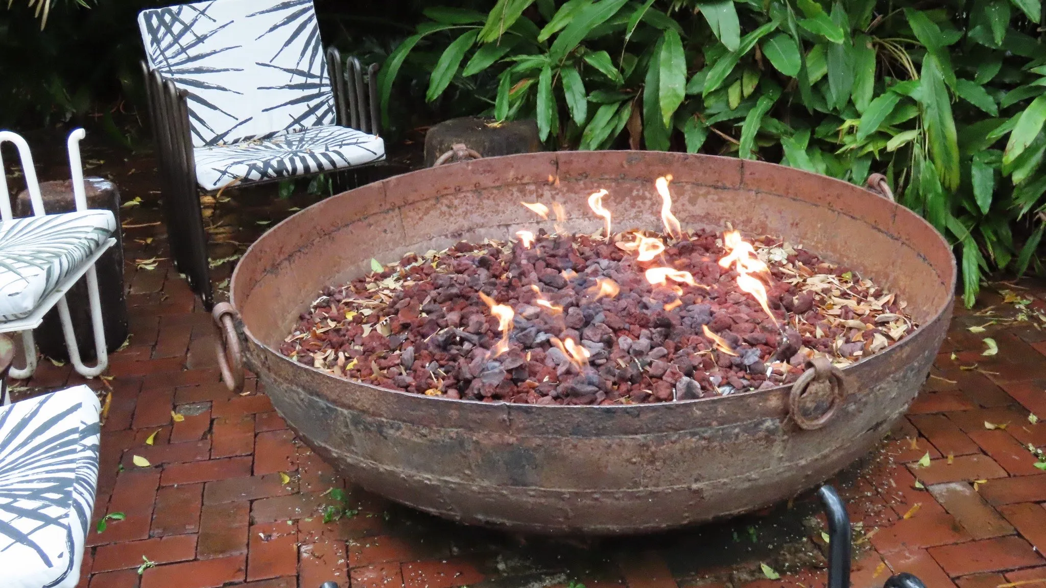 A wide steel gas fire bowl burning over lava rock on a brick backyard patio, patio chairs and tropical planting behind it