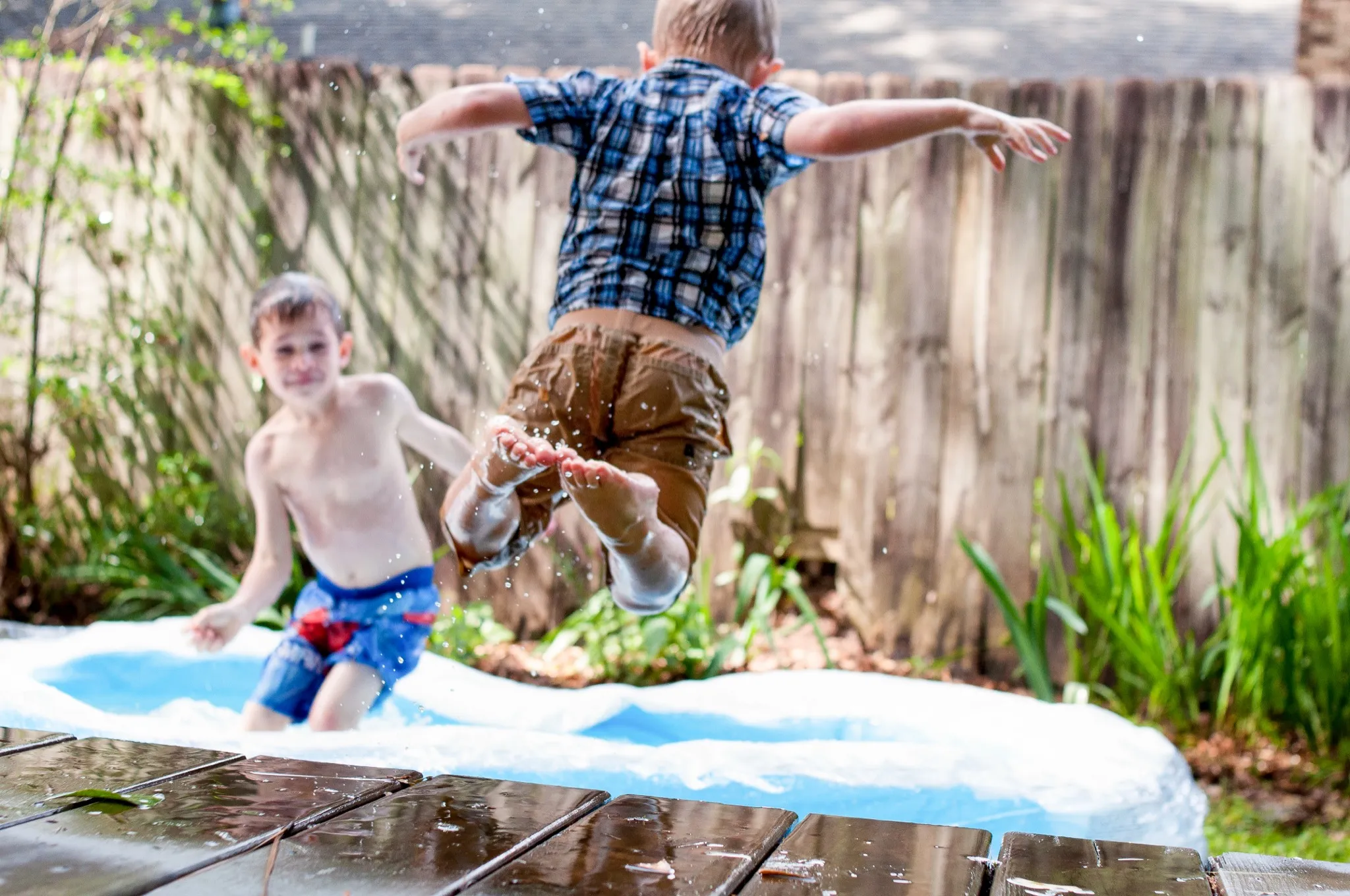Two kids splashing at a blue inflatable pool in a fenced backyard on a summer day