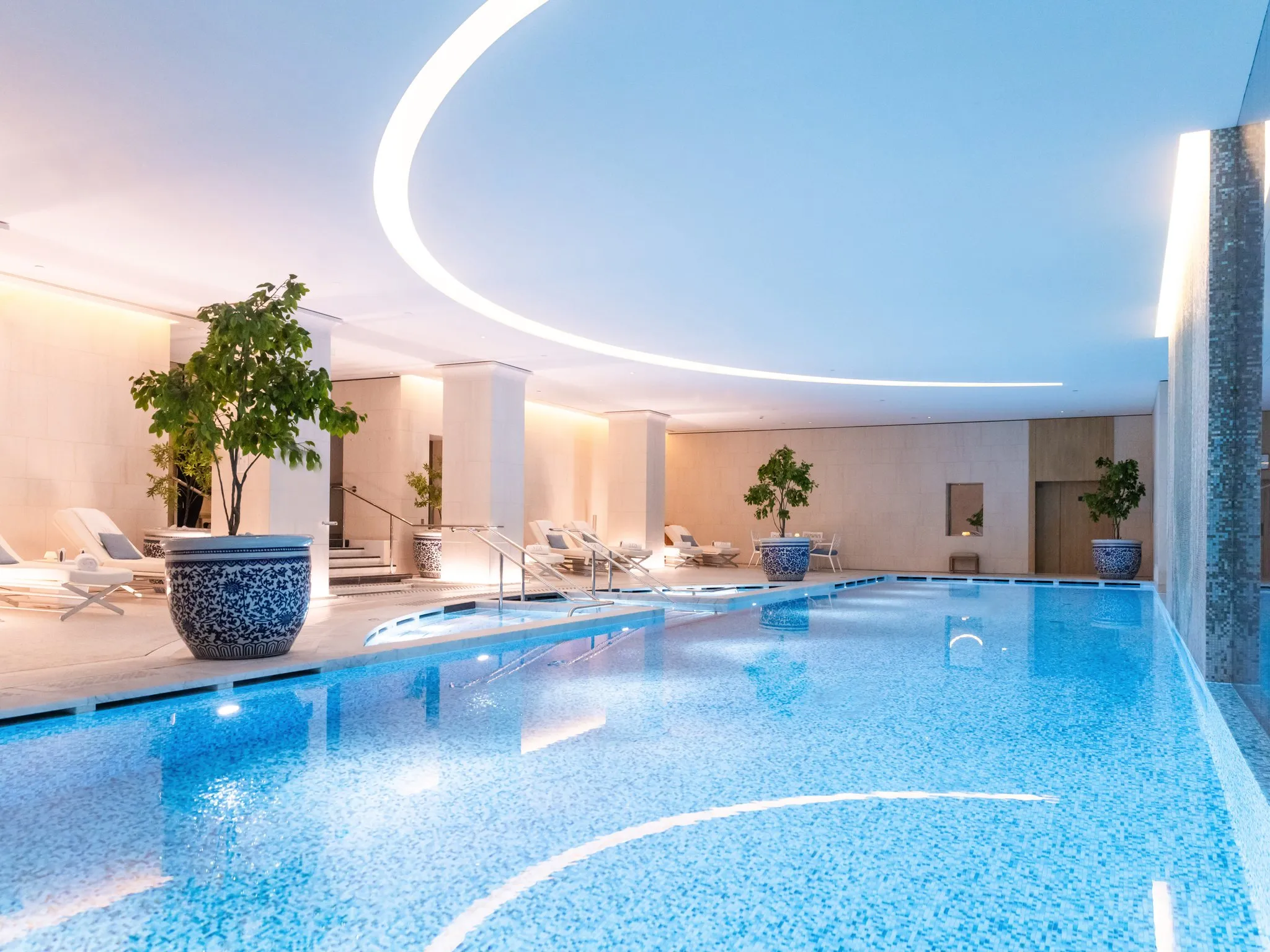 A clear blue resort swimming pool with a raised spa spilling into it and sun loungers along the deck