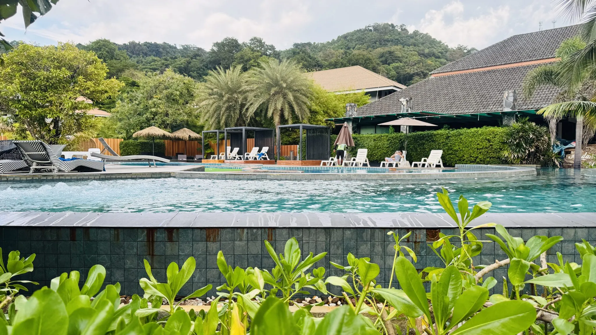 Wide view of a resort swimming pool ringed by white lounge chairs, cabanas and straw umbrellas under a blue sky