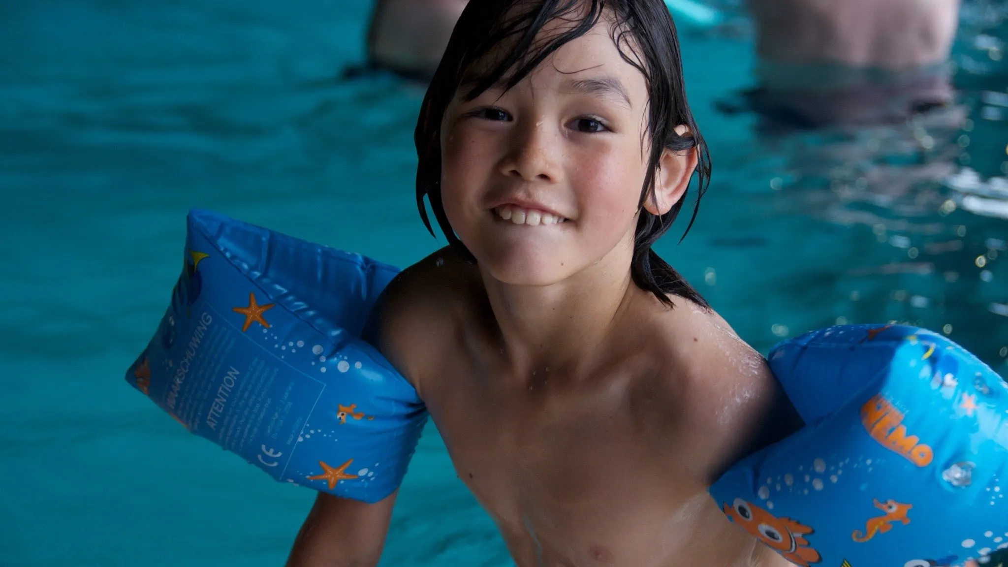 A young child in a swimming pool wearing inflatable armbands, seen across the water