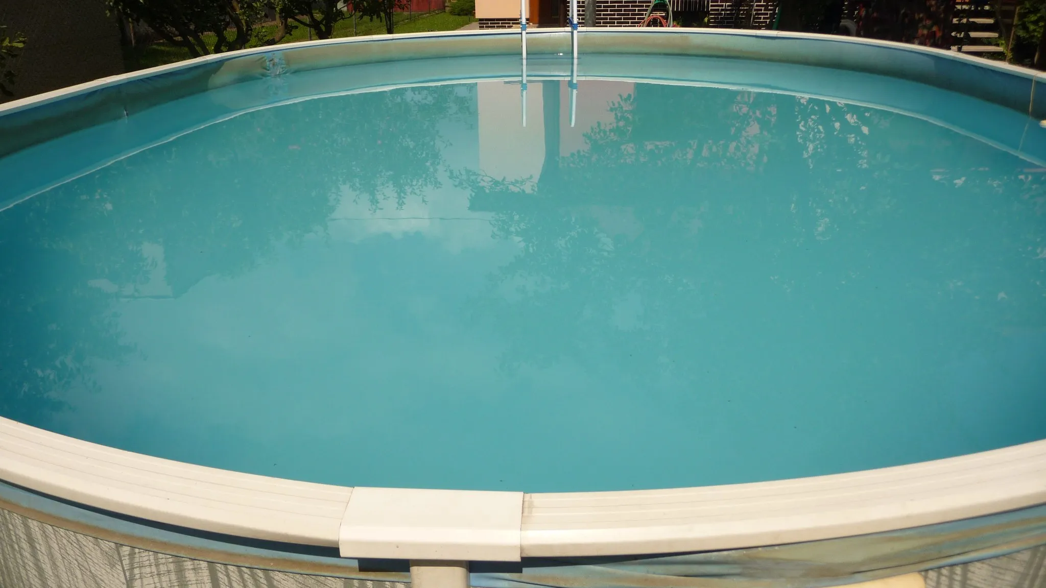 An oval above-ground pool seen across its water from the rim, with the ladder at the far end and the lawn behind