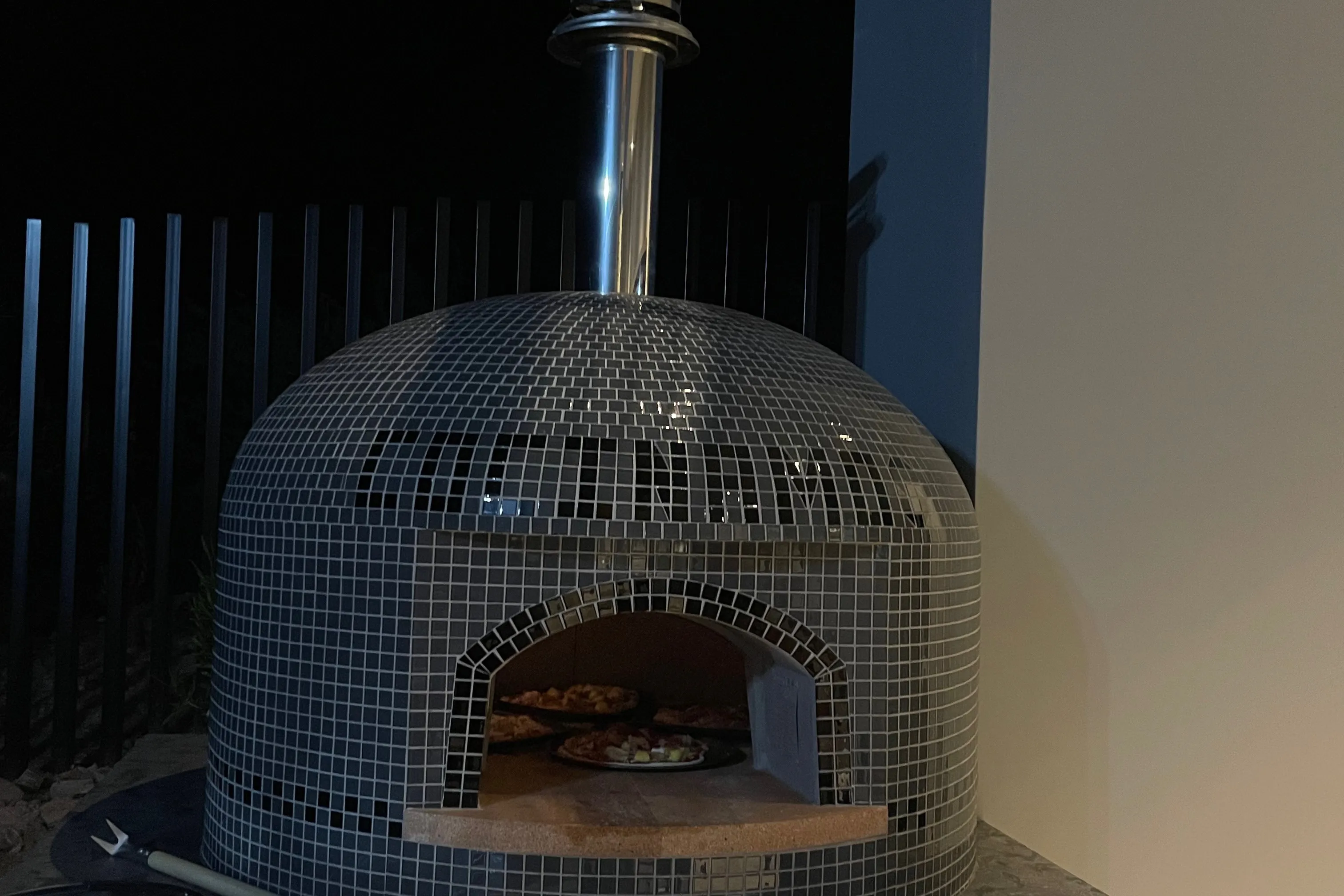 A tiled dome pizza oven glowing at night in an outdoor kitchen, with pizzas baking inside